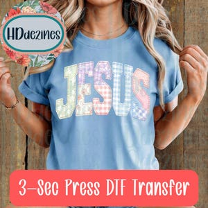 May include: Light blue t-shirt with the word "JESUS" in patchwork letters. The letters are in pastel colors and various patterns. The text "3-Sec Press DTF Transfer" is in a red rectangle at the bottom.