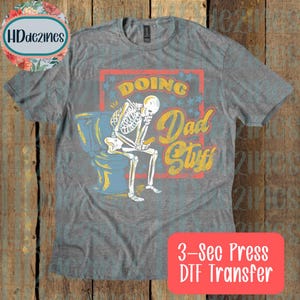 May include: A heather gray t-shirt featuring a graphic of a skeleton sitting on a toilet while looking at a phone. The text reads "Doing Dad Stuff" in yellow and white script. The design is set against a red rectangular background with blue stars.