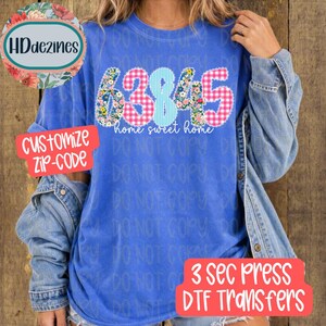 May include: A blue t-shirt featuring the text 63845 in a mix of floral and pink gingham patterns, with the words home sweet home written below in white script. The shirt is layered under a blue denim jacket with visible metal buttons on the sleeves.