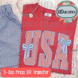 May include: Red t-shirt with the word "USA" in a patriotic design featuring red and white stripes, light blue bows, and a light blue outline. The shirt is paired with blue and white striped shorts and a silver necklace.