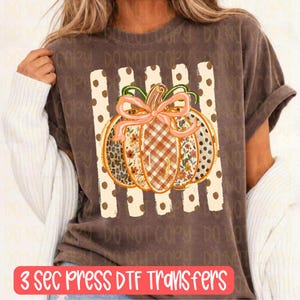 May include: A dark brown short-sleeve t-shirt featuring a central graphic of a pumpkin with a pink bow. The pumpkin has sections with leopard print, floral patterns, and plaid. The graphic is set against a background of vertical cream stripes with brown dots.