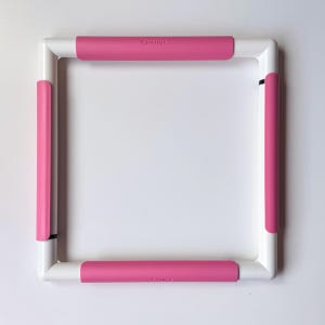 Op de afbeelding: Een vierkant borduurraam bestaande uit een witte kunststof structuur met vier roze kunststof clips aan elke zijde. De bovenste clip is voorzien van de tekst Q-Snap in reliëf en de onderste clip is voorzien van de tekst Q-Snap.