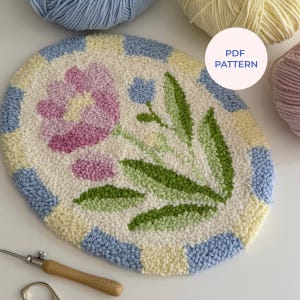 Op de afbeelding: Ovaal, handgetuft tapijt met een bloemenontwerp. Het tapijt heeft een centrale roze bloem, groene bladeren en een blauw en geel geblokte rand. De tekst "PDF PATTERN" staat in een witte cirkel. Garenkluwens staan op de achtergrond.