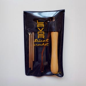 Pode incluir: Um conjunto de agulhas de crochê azul marinho em uma capa de vinil preta. O conjunto inclui uma agulha de crochê com cabo de madeira, três agulhas de crochê de metal e fio laranja. A capa tem a palavra "Lavor" e as palavras "tricot crochet" impressas em dourado.