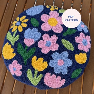 Puede incluir: Un cojín redondo azul marino con flores bordadas en rosa, amarillo y azul, con hojas verdes. El cojín presenta un patrón floral colorido y una etiqueta "PDF PATTERN".