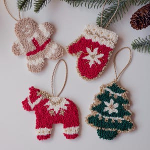 Op de afbeelding: Vier handgemaakte kerstversieringen: een peperkoekmannetje, een rode want, een rood paard en een groene kerstboom. Elk ornament is gemaakt van getextureerd garen, heeft gouden accenten en een lus om op te hangen.
