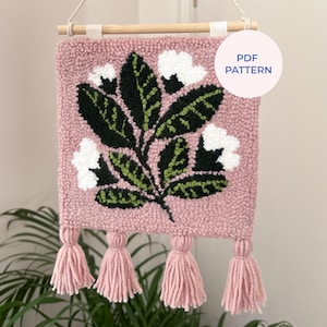 Puede incluir: Un tapiz de pared rosa texturizado con un diseño floral. El diseño presenta flores blancas y hojas verdes y negras. El tapiz tiene borlas rosas en la parte inferior y está suspendido de una clavija de madera. El texto "PDF PATTERN" está en la parte superior derecha.