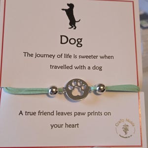 May include: A bracelet featuring a silver paw print charm and a light green cord. The card behind the bracelet has a dog silhouette and the word "Dog". The text on the card reads "A true friend leaves paw prints on your heart."