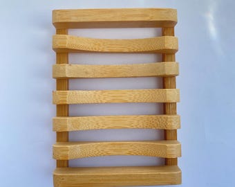 May include: A rectangular bamboo soap dish featuring a slatted design with six horizontal bars connected by two vertical supports. The light tan wood grain is visible across the surface, and the bars have a slightly curved, concave shape.