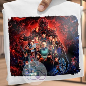 May include: White t-shirt featuring a vibrant, detailed graphic of characters from a popular science fiction series. The artwork showcases a group of people against a fiery red and dark blue background, with a monster figure prominently displayed.