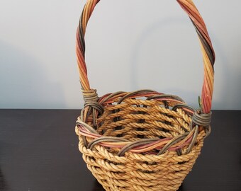 May include: A woven basket with a handle. The basket is made of natural-colored woven material with a round base and a handle that arches over the top. The handle is made of twisted strands in shades of orange, red, and brown.