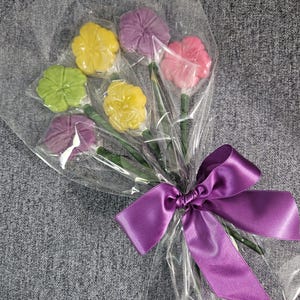 May include: A bouquet of colorful flower-shaped candies wrapped in clear cellophane. The candies are in shades of yellow, green, pink, and purple. The bouquet is tied with a purple satin ribbon.