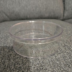 Könnte beinhalten: Ein runder Behälter aus klarem Kunststoff mit leicht ausgestelltem Boden. Der Behälter ist leer und steht auf einer grauen Stoffoberfläche.