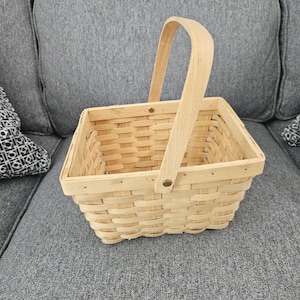 May include: A rectangular woven basket made of light-colored wood strips. The basket features a single arched handle attached to the sides with small metal rivets. The construction consists of horizontal and vertical woven slats.
