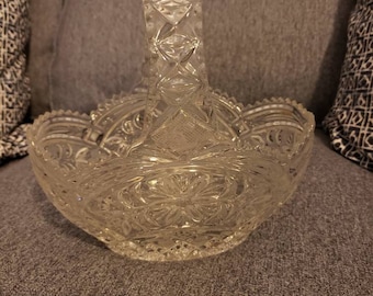 May include: A clear glass bowl with a decorative cut-glass pattern and a handle. The bowl is sitting on a gray fabric surface.