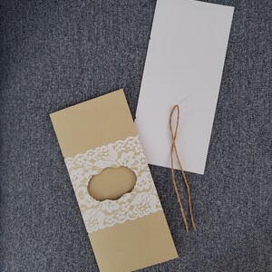 Può includere: Un biglietto d'invito beige con un motivo in pizzo bianco e una finestra fustellata. Sono presenti anche un biglietto bianco e un pezzo di spago. Gli elementi sono disposti su una superficie di tessuto grigio, adatti per annunci di matrimonio o eventi.
