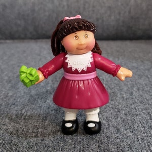 Puede incluir: Una figura de muñeca Cabbage Patch Kids vintage. La muñeca tiene cabello castaño, un lazo rosa y viste un vestido burdeos con cuello blanco. Sostiene un regalo verde. La muñeca lleva zapatos negros y calcetines blancos.