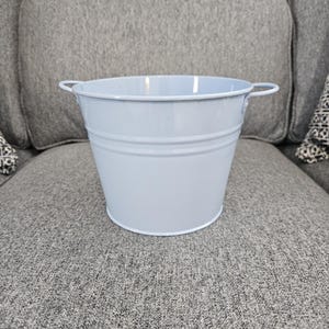 May include: A light blue metal bucket with two small handles. The bucket has a rounded shape with a white rim and two horizontal bands around the body. The bucket is sitting on a gray surface.