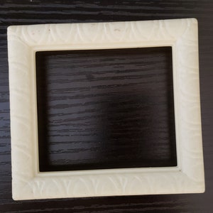 May include: A square, cream-coloured picture frame with a repeating pattern along the edges. The frame is empty, showing the dark surface beneath. The frame is made of a solid material.