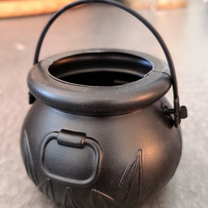 Puede incluir: Un caldero de plástico negro con asa, diseñado para el truco o trato de Halloween. El caldero tiene una superficie texturizada con diseños de hojas y una pequeña asa en el lateral.