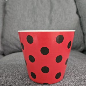 May include: A red flower pot with black polka dots. The pot has a white rim and is sitting on a gray surface. The pot is cylindrical and appears to be made of paper or cardboard.