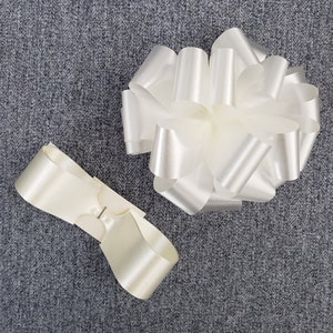 Może przedstawiać: Dwie kremowe satynowe kokardy na szarym tle. Jedna kokarda jest w pełni uformowana, a druga to płaska, wstępnie zawiązana kokarda z metalowym zapięciem. Kokardy są prawdopodobnie przeznaczone do pakowania prezentów lub dekoracji.