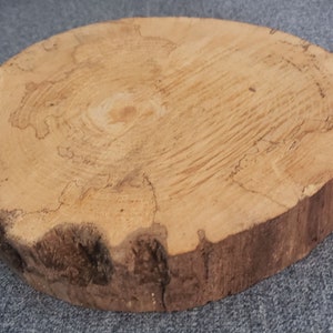 May include: A round wooden slice with a natural wood grain pattern and a rough, uneven edge. The wood is a light brown color with darker brown streaks.