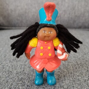 Puede incluir: Muñeca vintage de Cabbage Patch Kids con uniforme de banda de música. La muñeca tiene pelo negro de hilo, ojos marrones y tez clara. Lleva una chaqueta roja con charreteras doradas, un sombrero azul con una pluma roja y botas azules. La muñeca sostiene un bastón de caramelo.