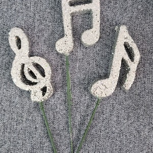 Peut inclure: Trois pics de notes de musique argentées à paillettes. Les pics sont en forme de clé de sol, de noire et de croche. Les pics sont sur un fond gris.
