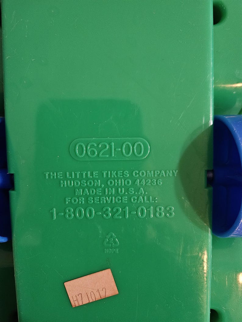 Puede incluir: Primer plano de un componente de juguete de pl&aacute;stico verde con el texto "0621-00". Tambi&eacute;n se ve el texto "THE LITTLE TIKES COMPANY" y un n&uacute;mero de tel&eacute;fono. Una peque&ntilde;a pegatina con el texto "H7J012" est&aacute; adjunta.