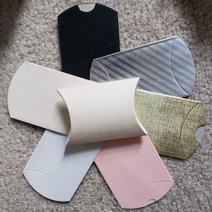 Könnte beinhalten: Eine Sammlung kissenförmiger Geschenkboxen in verschiedenen Farben, darunter Schwarz, Creme, Silber mit diagonalen Streifen, Gold, Hellrosa und Weiß. Die Boxen bestehen aus strukturiertem Papier und liegen auf einem neutralfarbenen Teppich.