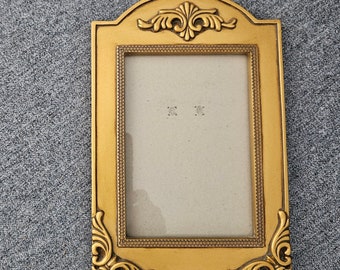 Op de afbeelding: Een goudkleurige fotolijst met sierlijke details. Het frame heeft een rechthoekige opening met een decoratieve rand en een gebogen bovenkant. De hoeken en de bovenkant hebben krullende versieringen. Het frame is leeg.