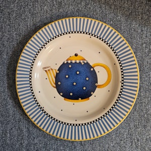 Pode incluir: Um prato de cerâmica decorativo com um desenho de bule azul. O prato tem um fundo branco com uma borda listrada azul e branca. O bule é azul com bolinhas brancas e detalhes amarelos.