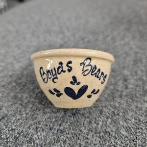 Puede incluir: Pequeño cuenco de cerámica beige con la inscripción "Boyds Bears" en escritura azul oscuro. El cuenco presenta un diseño de corazón y hojas en azul oscuro. Es probable que sea un artículo de colección o decorativo.