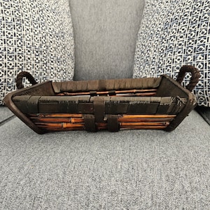 May include: A rectangular brown bamboo basket with dark brown woven accents and rope handles. The basket has a dark brown rim and is sitting on a gray surface.