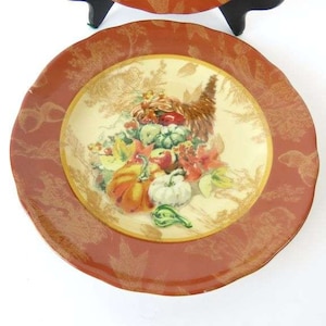 Puede incluir: Un plato decorativo con un tema de cosecha de otoño. El plato presenta una imagen central de calabazas, calabacines y otros productos de otoño. El borde es de color marrón rojizo con un diseño dorado. El plato probablemente se utiliza para servir alimentos o como artículo decorativo.