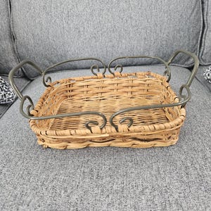 May include: A rectangular woven wicker basket featuring a light tan natural finish. The basket is framed by a dark gray metal structure with curved, scroll-like handles on each side, providing a sturdy border around the woven base.
