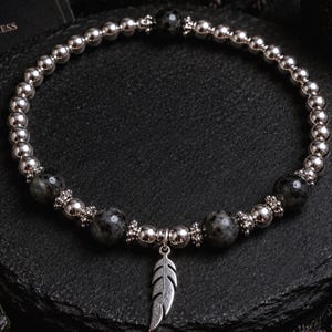 May include: A beaded bracelet featuring alternating polished silver-tone metallic spheres and dark grey speckled stone beads. The design includes small decorative silver-tone spacers and a central silver-tone metal feather charm pendant.