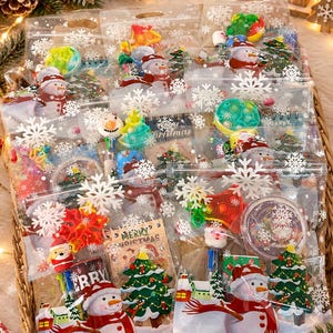 Op de afbeelding: Transparante plastic cadeauzakjes versierd met witte sneeuwvlokken, elk met een kleine speelgoedset in kerstthema. De sets bevatten items zoals mininotitieboekjes, pennen met figuurtjes en kleurrijke siliconen fidget-speeltjes.