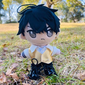 Pode incluir: Boneca de pelúcia de um personagem com cabelo preto, usando um colete dourado, camisa branca e sapatos de verniz pretos. A boneca tem olhos grandes e está em uma área gramada com um fundo desfocado de árvores.