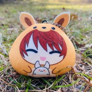 May include: A plush keychain featuring a cartoon character in a bunny costume. The keychain is primarily orange with brown accents, including the ears and hair. The character has a smiling face and a small bunny graphic on the front.