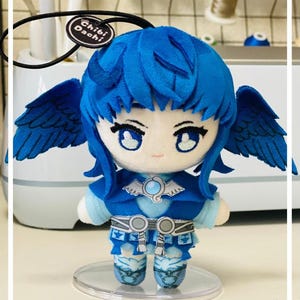Puede incluir: Muñeca de peluche con pelo, alas y ropa azules. La muñeca tiene ojos grandes y está de pie sobre una base de plástico transparente. El atuendo de la muñeca incluye una parte superior azul con un emblema plateado y botas a juego. La etiqueta dice "Chibi Dachi".