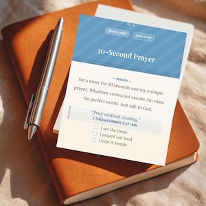 May include: A silver pen lies on a closed, brown leather journal, with a white card on top. The card has a blue header with the text "30-Second Prayer" and instructions to pray for 30 seconds. The journal and card are on a textured surface.