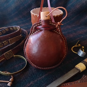 Peut inclure: Une gourde en cuir marron avec un bouchon en bois et des coutures décoratives. La gourde est dotée d'une lanière en cuir et est entourée d'autres accessoires en cuir, notamment des ceintures et une petite pochette. Les articles sont présentés sur un tissu à motifs.