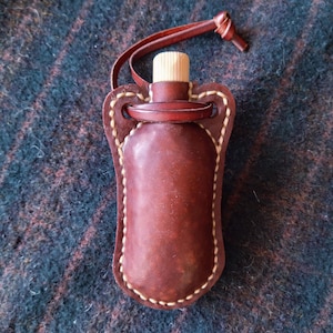 May include: A small, brown leather flask with a wooden stopper and a leather loop. The flask is encased in a stitched leather holder with a decorative design. The background is a textured, dark fabric.
