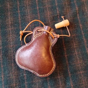 May include: A brown leather flask with a light brown stitched border and a wooden stopper. The flask has a teardrop shape and is connected to the stopper with a thin, light brown leather cord. The flask is empty.
