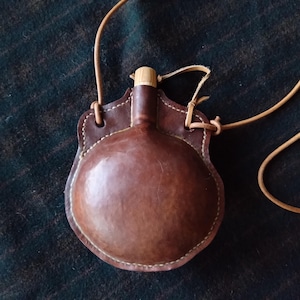 May include: A brown leather flask with a wooden stopper and a leather strap. The flask is encased in a stitched leather holder with a rounded shape. The design is rustic and the leather has a natural, aged appearance.