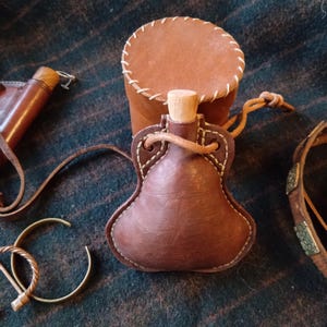 May include: A selection of brown leather items, including a flask with a wooden stopper, a pouch, and a bracelet. The flask is stitched with white thread and has a leather cord. The items are displayed on a textured, dark green surface, reminiscent of a traditional textile.