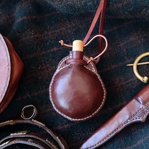 May include: A small, brown leather flask with a wooden stopper and a stitched leather casing. The flask is suspended by a thin, brown leather cord. Other leather accessories are visible in the background.