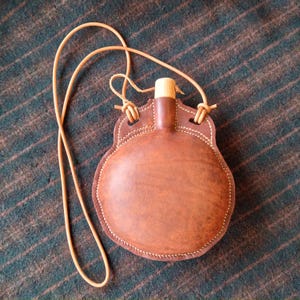 May include: A brown leather flask with a light brown leather strap. The flask has a rounded body and a wooden stopper. The edges are stitched with white thread. The flask is designed to be worn around the neck.